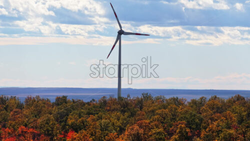 Video - Aerial drone view of a wind turbine rising above colorful autumn forest in Moldova under a bright blue sky