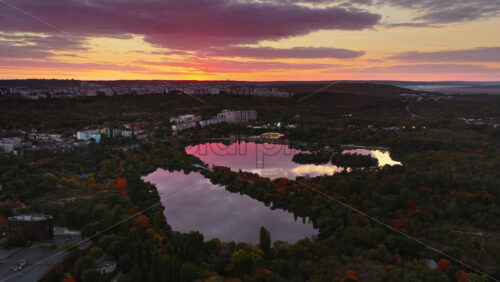 Video - Aerial drone view of a lake reflecting the colorful evening sky and surrounding cityscape, capturing a peaceful urban moment with autumn foliage in Chisinau, Moldova, at sunset