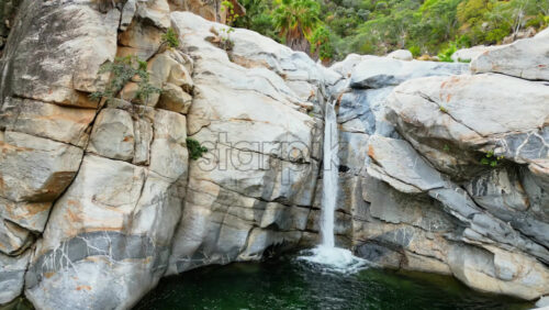 Video - Aerial drone view of a small waterfall cascading between rocks surrounded by desert vegetation and palm trees in Baja California Sur, Mexico