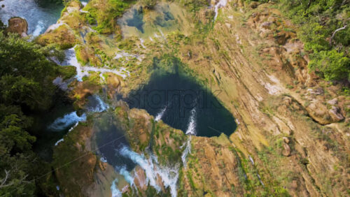 Video - Aerial drone view showing a waterfall flowing into a deep blue basin surrounded by rock formations and vegetation
