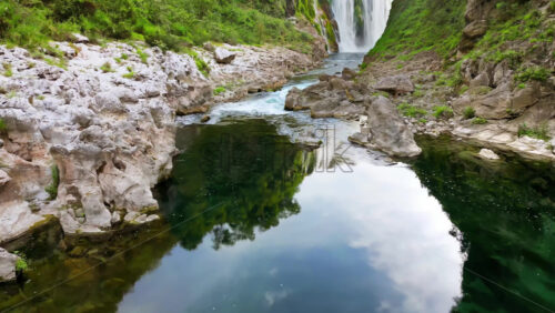 Video - Aerial drone view a waterfall plunging into a crystal clear pool, surrounded by rocky cliffs and lush greenery