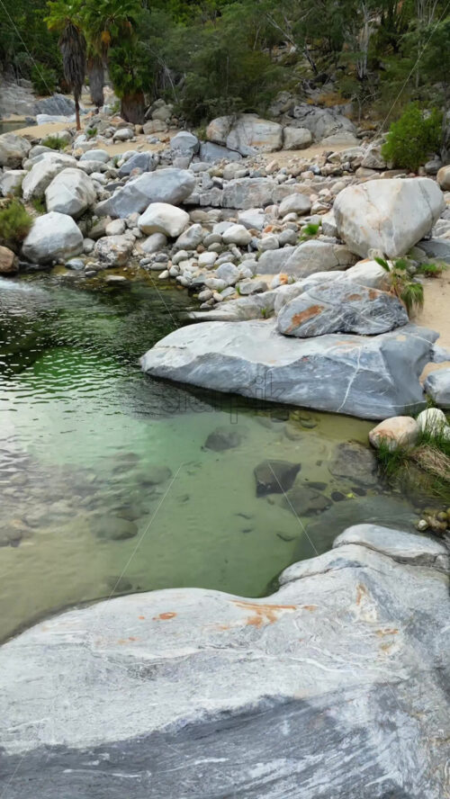 Video - Aerial drone view of a small waterfall cascading between massive rocks into a natural pool in Baja California Sur's desert canyons. Vertical