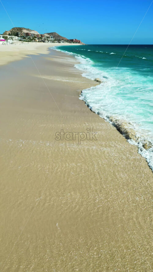 Video - Aerial drone view of turquoise waves gently washing over golden sand under a clear blue sky at Costa Azul Beach, Los Cabos. Vertical