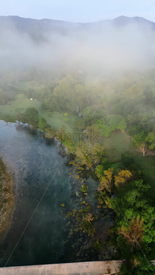 Video - Aerial drone view of a calm river flowing under a bridge surrounded by dense vegetation and morning fog. Vertical