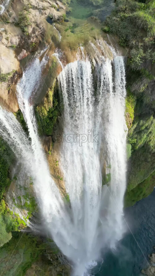 Video - Aerial drone view of Tamul Waterfall cascading down green cliffs into a turquoise pool below. Vertical