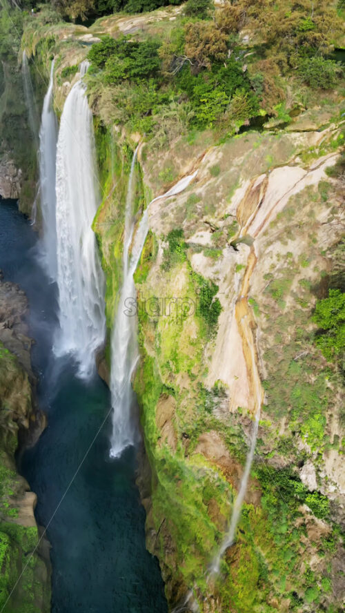 Video - Aerial drone view of Tamul Waterfall cascading down green cliffs into a turquoise pool below. Vertical
