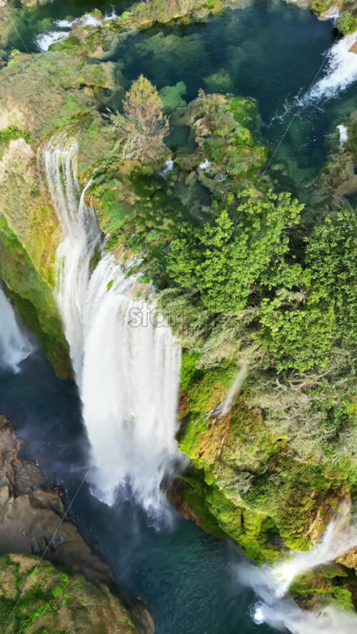 Video - Aerial drone view of Tamul Waterfall cascading down green cliffs into a turquoise pool below. Vertical