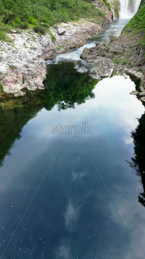 Video - Aerial drone view a waterfall plunging into a crystal clear pool, surrounded by rocky cliffs and lush greenery. Vertical