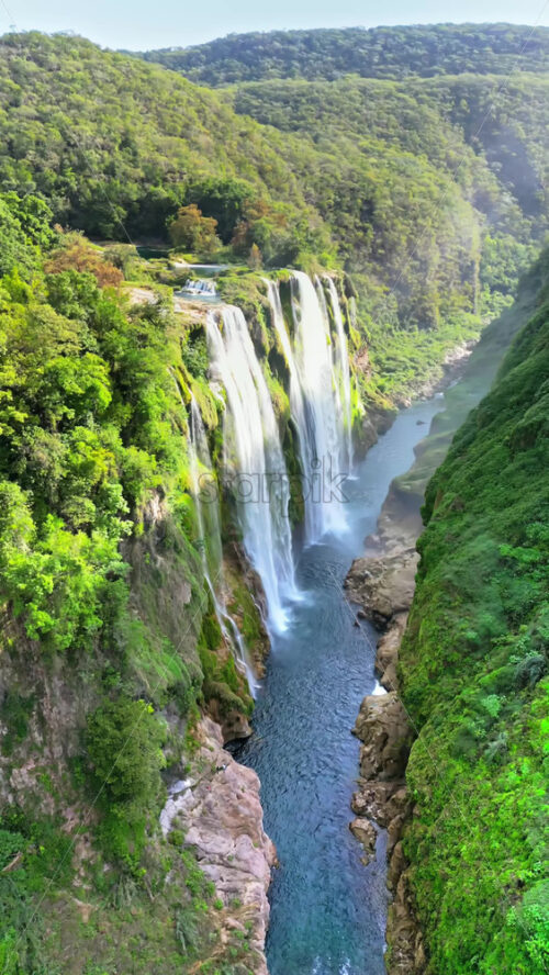 Video - Aerial drone view of Tamul Waterfall cascading down green cliffs into a turquoise pool below. Vertical