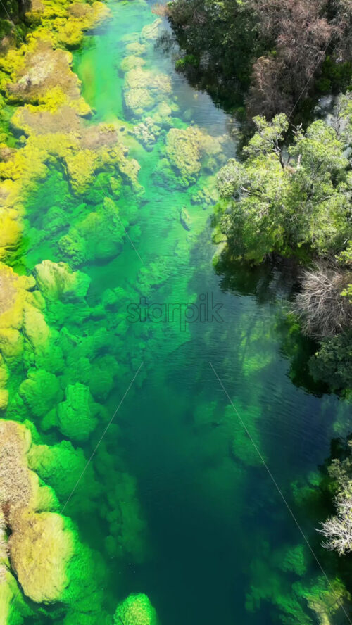 Video - Aerial drone view showing a waterfall flowing into a deep blue basin surrounded by rock formations and vegetation. Vertical