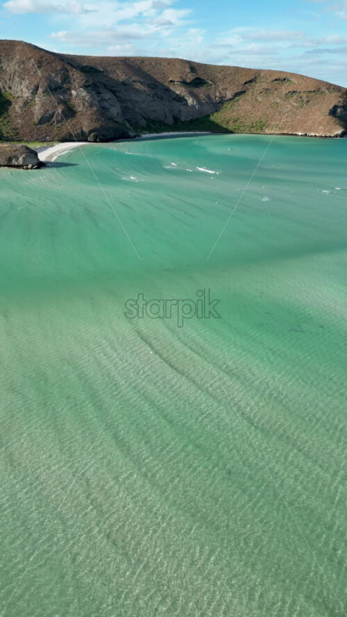 Video - Aerial drone view of turquoise sea waters reflecting the afternoon sun, surrounded by arid mountains in Baja California Sur. Vertical