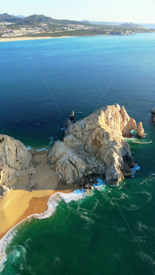 Video - Aerial drone view of Lover's Beach, one of the most famous beaches in Cabo San Lucas, framed by monumental rock cliffs and emerald waters. Vertical