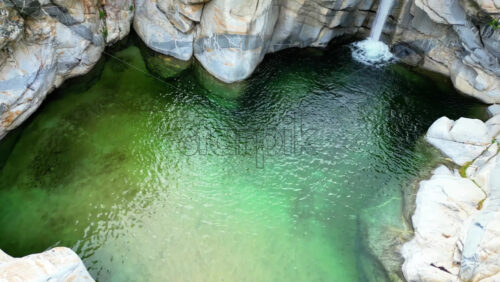 Video - Aerial drone view of a small waterfall cascading over smooth rocks, surrounded by palm trees and wild plants in slow motion