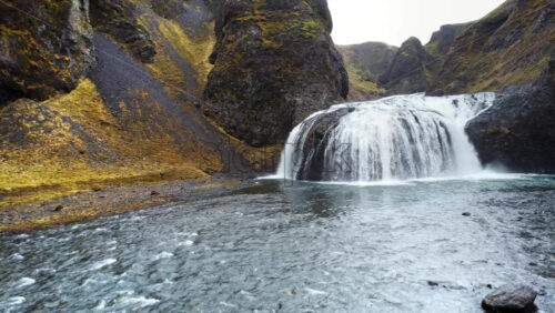 Video - Aerial drone view of a stunning Icelandic waterfall flowing into a clear blue river surrounded by dark volcanic cliffs and moss covered hills