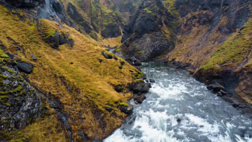 Video - Aerial drone view of a fast flowing river running between rugged cliffs and mossy volcanic rocks