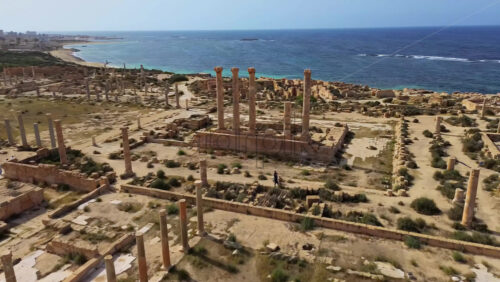 Video - Aerial drone view over the ancient ruins of Sabratha, Libya, showing remains of temples, streets, and columns surrounded by desert vegetation and bordered by the deep blue Mediterranean waters