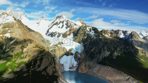 Video - Aerial drone view of a bright turquoise glacial lake nestled between steep rocky cliffs and snow covered peaks