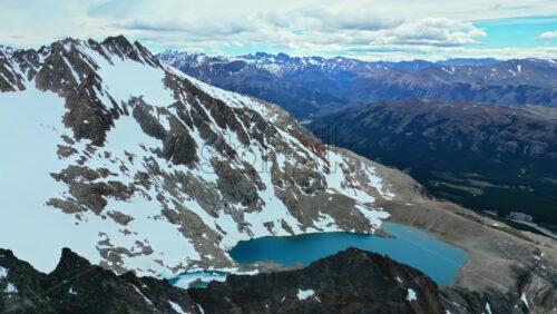 Video - Aerial drone view of a bright turquoise glacial lake nestled between steep rocky cliffs and snow covered peaks
