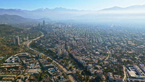 Video - Aerial drone view of the dense urban sprawl of Santiago, Chile, surrounded by distant Andes peaks under a hazy morning sky.