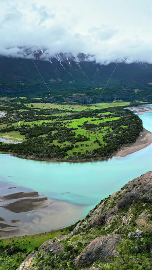 Video - Aerial drone view of a serene Patagonian landscape featuring a winding river, green plains, and mountains partially covered by clouds. Vertical