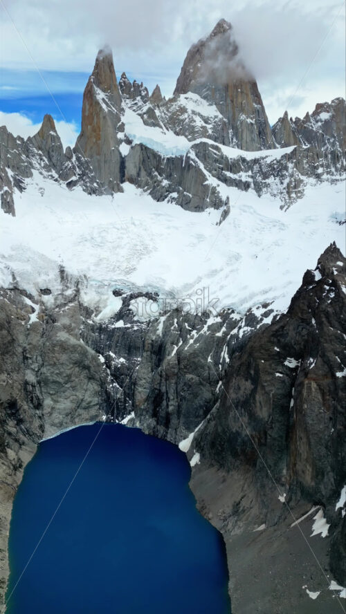 Video - Aerial drone view of the majestic peaks of Mount Fitz Roy rising above a deep blue glacial lake surrounded by rocky cliffs. Vertical