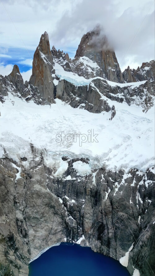 Video - Aerial drone view of the majestic peaks of Mount Fitz Roy rising above a deep blue glacial lake surrounded by rocky cliffs. Vertical