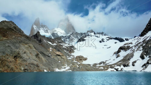 Video - Aerial drone view of a turquoise mountain lake surrounded by rugged terrain and snowfields beneath the Fitz Roy massif