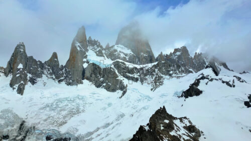 Video - Aerial drone view of the dramatic granite peaks of Fitz Roy partially covered by clouds with glaciers stretching across the slopes