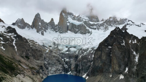 Video - Aerial drone view of the dramatic granite peaks of Fitz Roy partially covered by clouds with glaciers stretching across the slopes