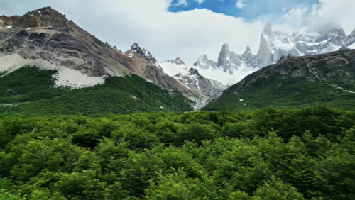 Video - Aerial drone view above lush green forest toward the towering peaks of Fitz Roy covered in snow and clouds
