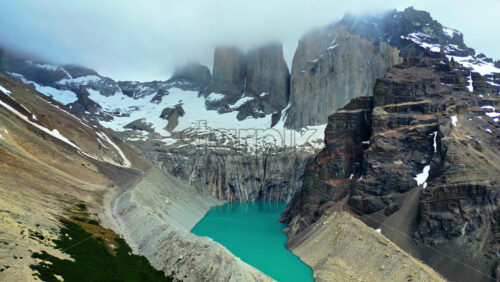 Video - Aerial drone view of the iconic Torres del Paine towers rising above a turquoise lake surrounded by snow and steep cliffs