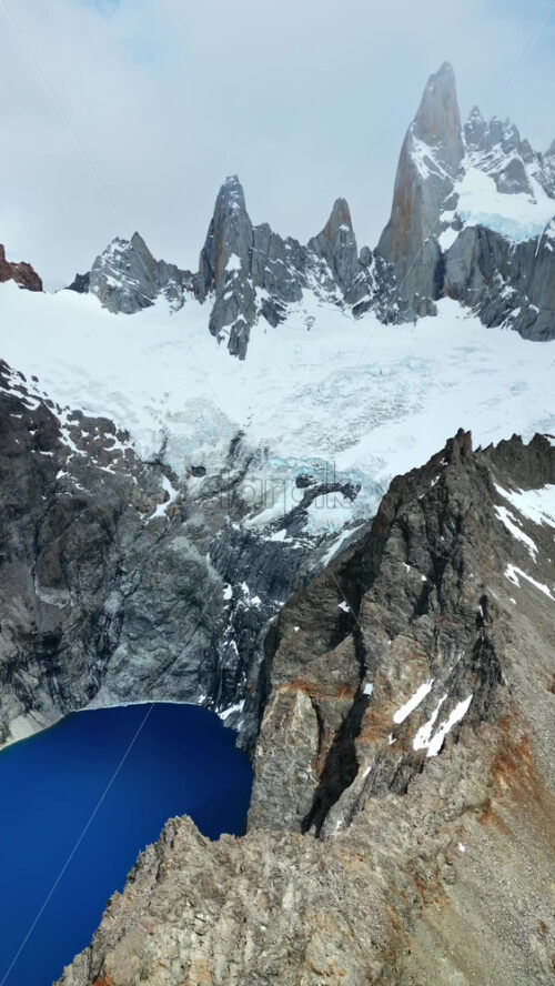 Video - Aerial drone view of the towering Fitz Roy peaks above a dark blue glacial lake framed by rocky cliffs and snow patches. Vertical