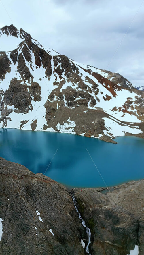 Video - Aerial drone view of a turquoise mountain lake surrounded by rugged terrain and snowfields beneath the Fitz Roy massif. Vertical