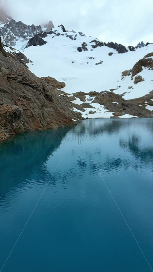 Video - Aerial drone view of a turquoise mountain lake surrounded by rugged terrain and snowfields beneath the Fitz Roy massif. Vertical