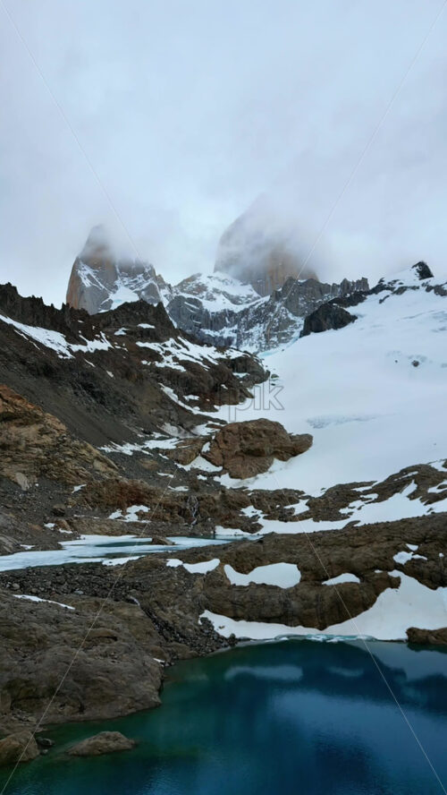 Video - Aerial drone view of a turquoise mountain lake surrounded by rugged terrain and snowfields beneath the Fitz Roy massif. Vertical