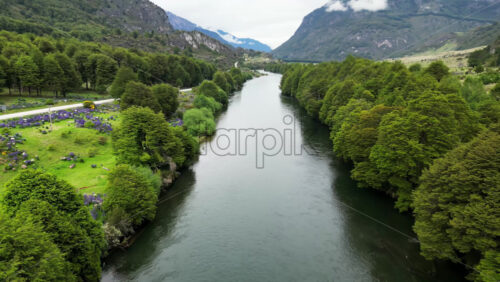 Video - Aerial drone view of a rustic wooden suspension bridge crossing a scenic river surrounded by trees and wildflowers