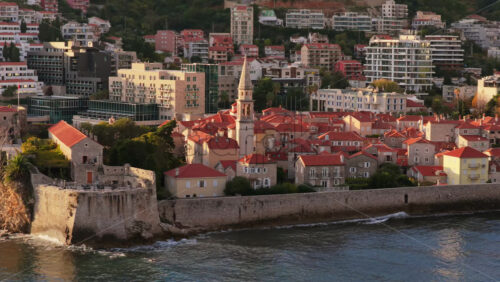 Video – Aerial drone view of Budva Old Town, showing its medieval stone buildings, red rooftops, fortress walls, and church tower along the coastline, with modern city buildings and mountains in the background. Montenegro - Starpik Stock