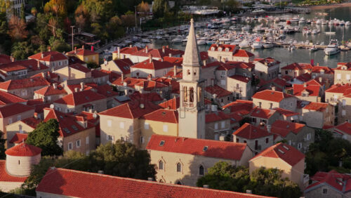 Video – Aerial drone view of Budva Old Town, showing its medieval stone buildings, red rooftops, fortress walls, and church tower along the coastline, with modern city buildings and mountains in the background. Montenegro - Starpik Stock