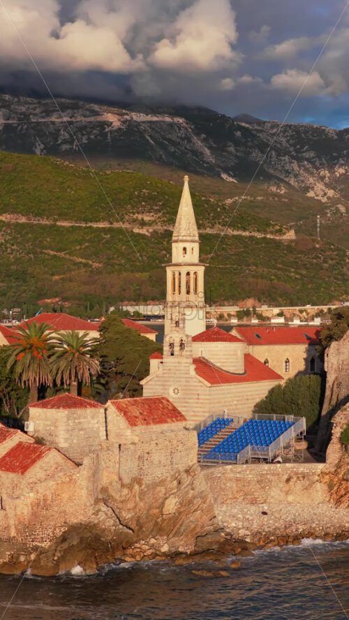 Video – Aerial drone view of Budva Old Town, showing its medieval stone buildings, red rooftops, fortress walls, and church tower along the coastline, with modern city buildings and mountains in the background. Vertical - Starpik Stock