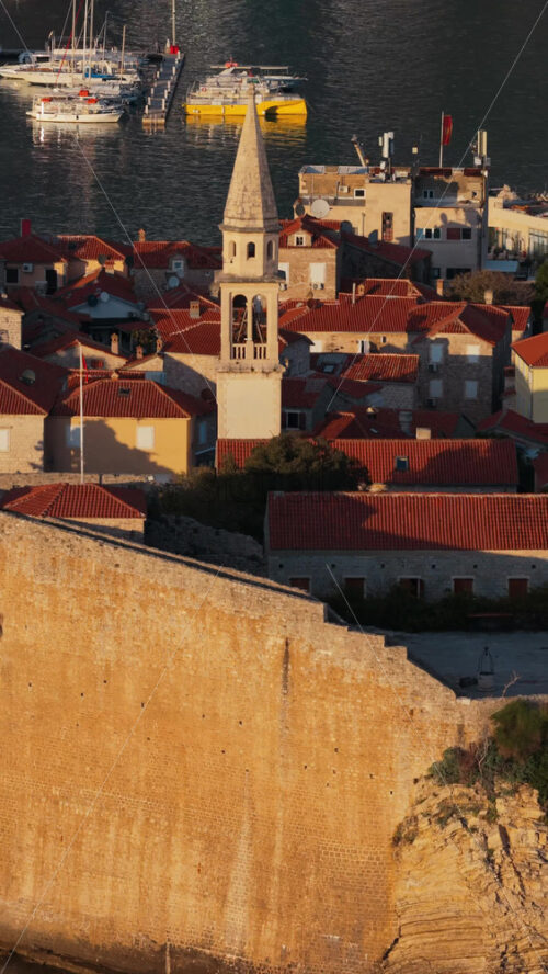 Video – Aerial drone view of Budva Old Town, showing its medieval stone buildings, red rooftops, fortress walls, and church tower along the coastline, with modern city buildings and mountains in the background. Vertical - Starpik Stock