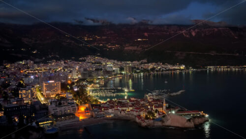 Video – Aerial drone view of Budva after sunset, featuring stone houses with red roofs and a tall church bell tower warmly illuminated from within. Montenegro - Starpik Stock