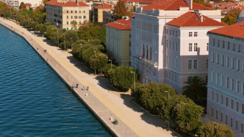 Video – Aerial drone view of Zadar’s historic seafront with bright Adriatic waters, elegant stone buildings, red rooftops, palm trees, and people walking along the promenade - Starpik Stock
