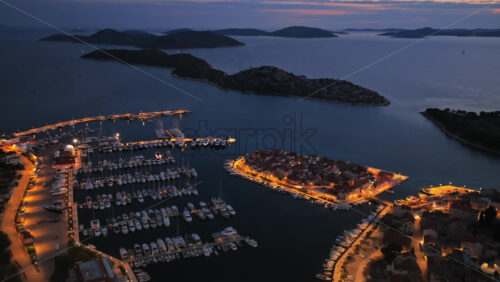 Video – Aerial drone view of a glowing marina filled with boats surrounding a compact old town of Tribunj Island illuminated by warm streetlights - Starpik Stock