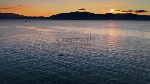 Video – Aerial drone view of a lone fisherman rowing his small wooden boat during sunset in Vlora Bay, Albania, with soft orange skies, calm waters, and distant silhouettes of hills and clouds - Starpik Stock