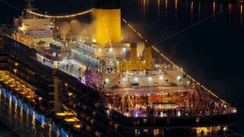 Video – Aerial drone view of a massive cruise ship, brightly lit with warm lights, as passengers enjoy its outdoor decks at night - Starpik Stock