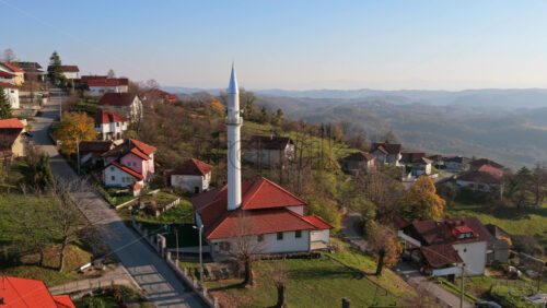 Video – Aerial drone view of a picturesque hillside village featuring red roofed houses and a tall white mosque minaret, surrounded by open fields and distant mountain ranges in Bosnia and Herzegovina - Starpik Stock