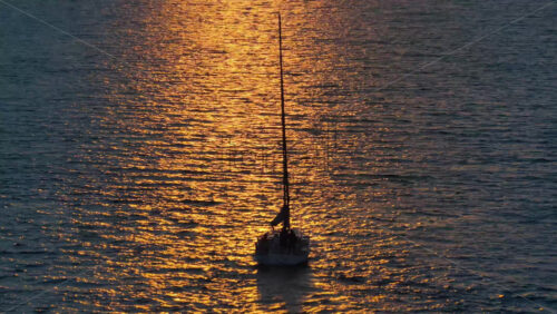 Video – Aerial drone view of a sailboat gliding on golden, sunlit water during sunset, with shimmering reflections surrounding the vessel - Starpik Stock