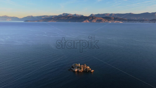 Video – Aerial drone view of an isolated rocky Peristeres Island featuring the old lighthouse ruins, illuminated by golden hour sunlight against the deep blue Adriatic Sea. Greece - Starpik Stock