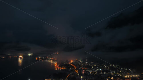 Video – Aerial drone view of the Bay of Kotor at night, with fog drifting across the landscape and a brightly lit cruise ship reflecting on the water. Coastlines glow with orange lights - Starpik Stock