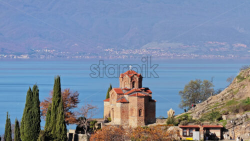 Video – Aerial drone view of the Church of St. John at Kaneo, surrounded by rocky cliffs, autumn trees, and small boats docked along the shoreline - Starpik Stock